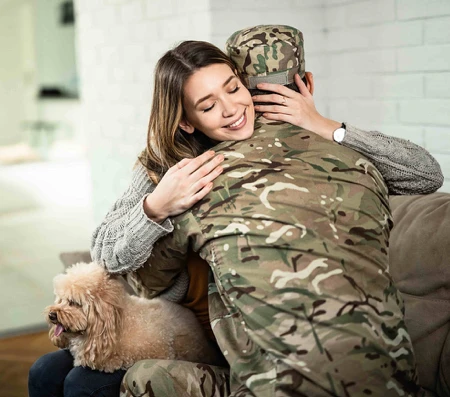 Person in military uniform embraced by a smiling woman in a home setting, with a small dog sitting beside them on the couch.
